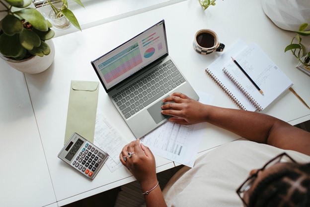 A woman sitting at a white table with two notebooks, a calculator, a small envelope, and a laptop working to streamline business processes.